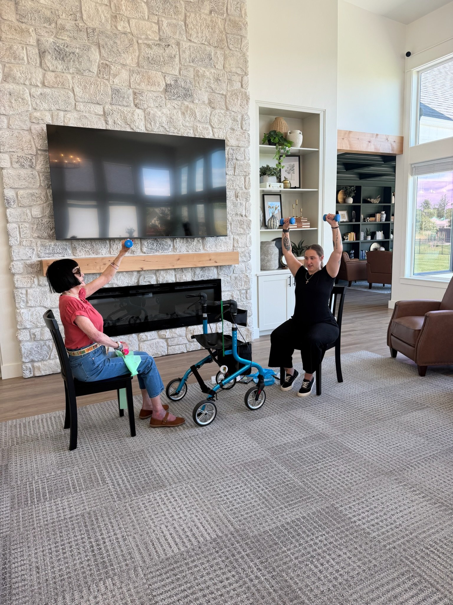 A resident and caregiver doing a seated dumbbell exercise class by the stone fireplace at The Ponderosa Estate