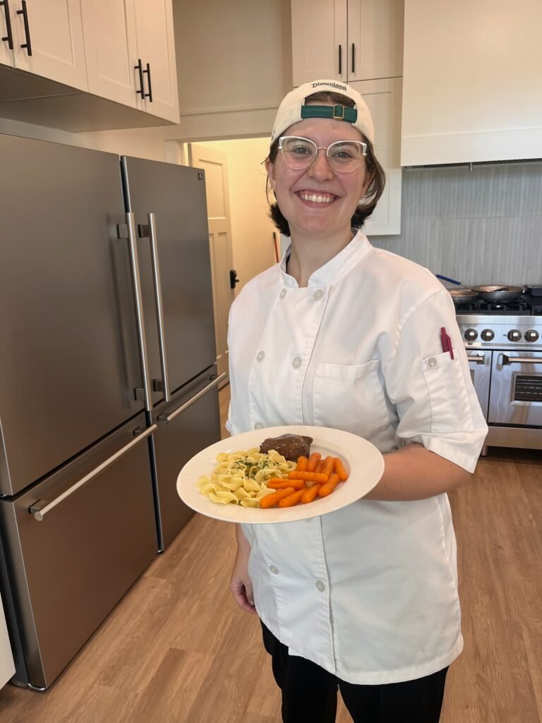 A smiling caregiver in a chef coat holding a plated meal in the kitchen at The Ponderosa Estate
