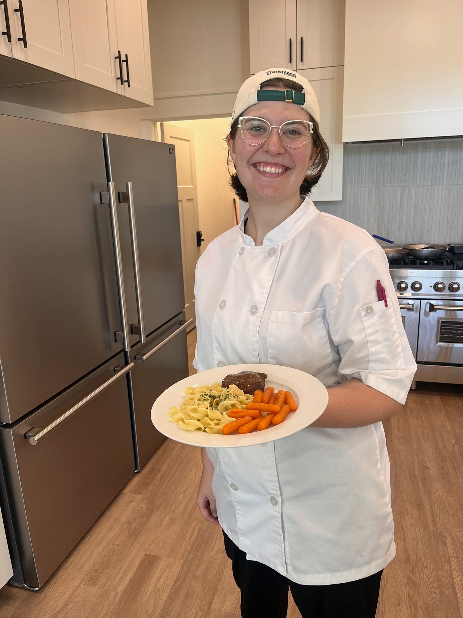 A smiling caregiver in a chef coat holding a plated meal in the kitchen at The Ponderosa Estate