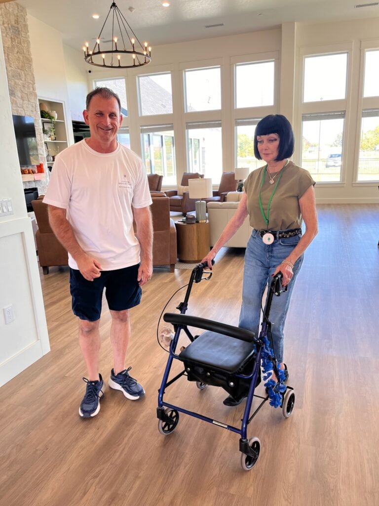 A Resident walking with her rollator alongside a smiling team member in the sunlit great room at The Ponderosa Estate in Eagle, Idaho